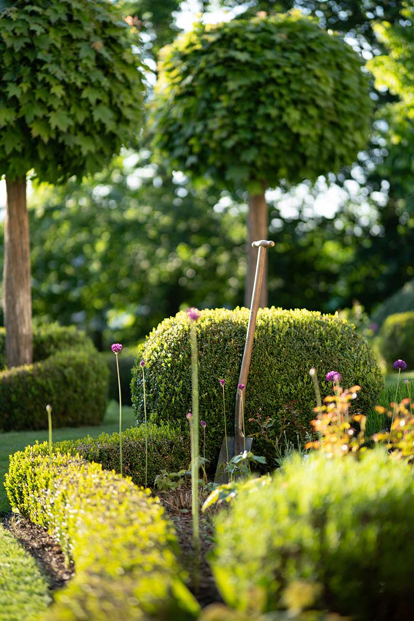 Manicured Garden With Rounded Topiary Shrubs, Flowering Plants, And A Garden Spade Resting Among The Landscaping Beds